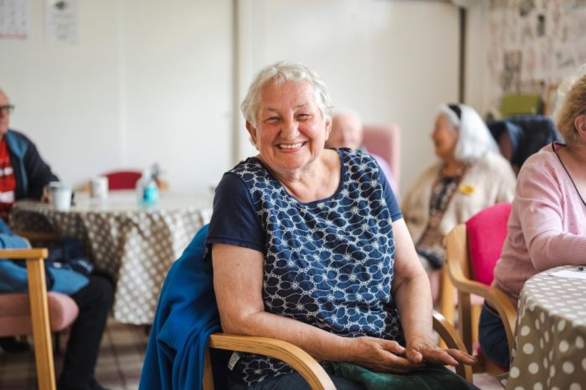 Eldre dame sitter på en stol og smiler til kamera