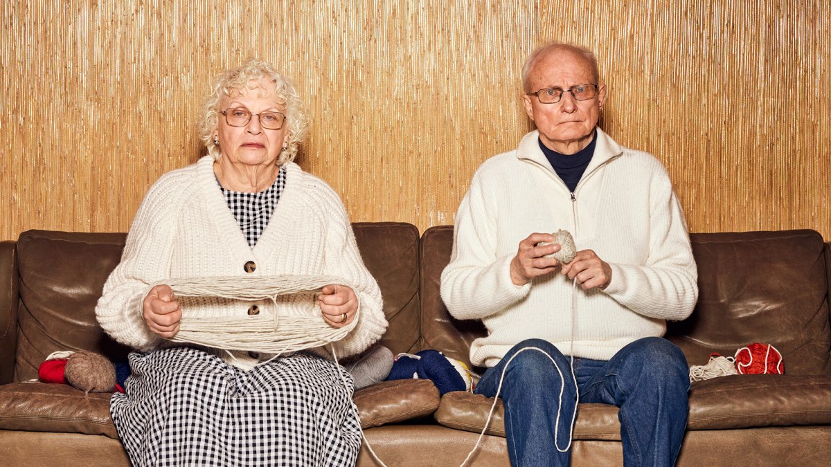 En gammel mann og en gammel dame sitter i en brun skinnsofa. Damen har tråd rundt hendene mens mannen nøster tråden. 